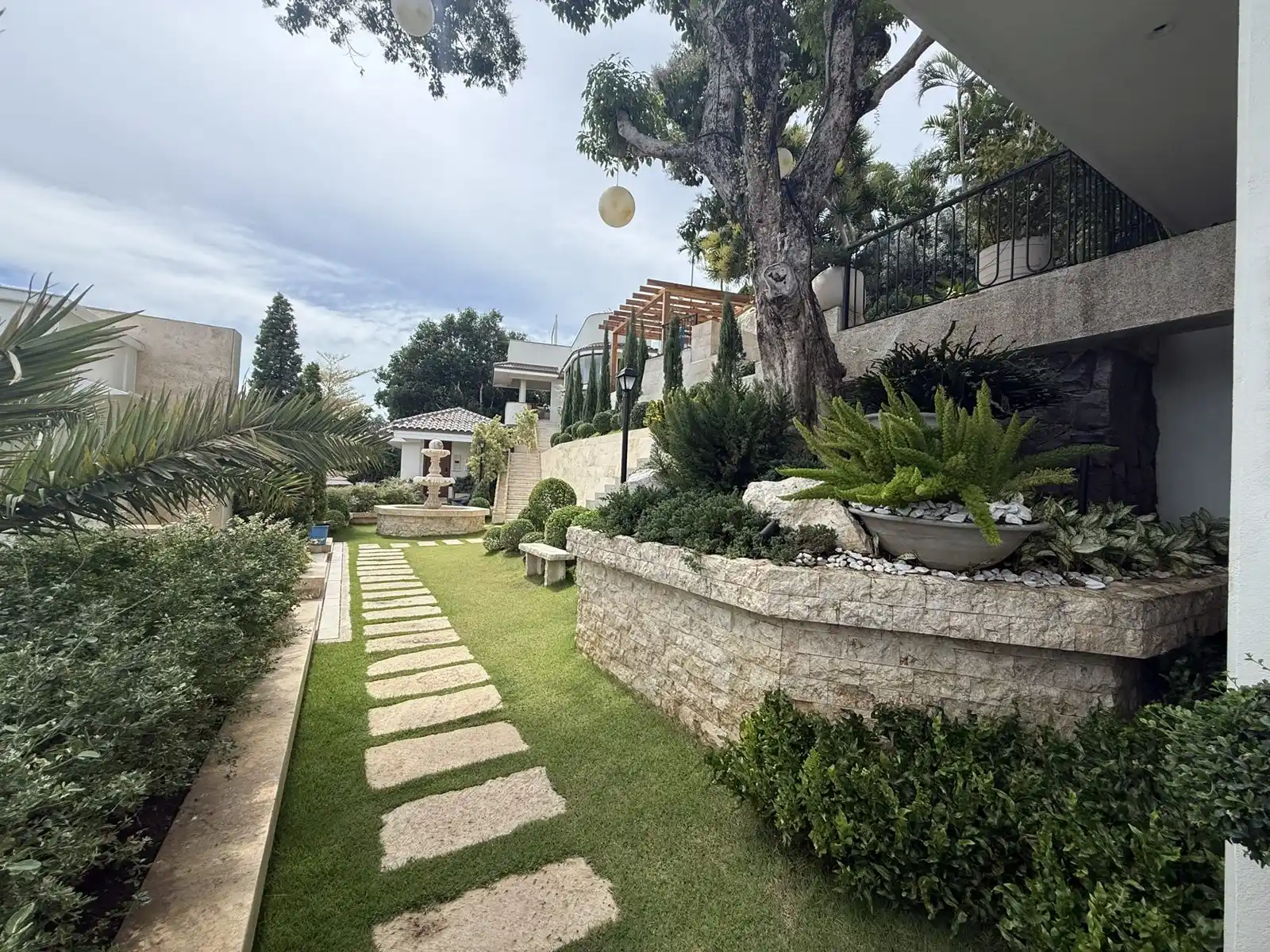 Stepping-stone garden path leading to a stone fountain, with layered planting and a mature feature tree