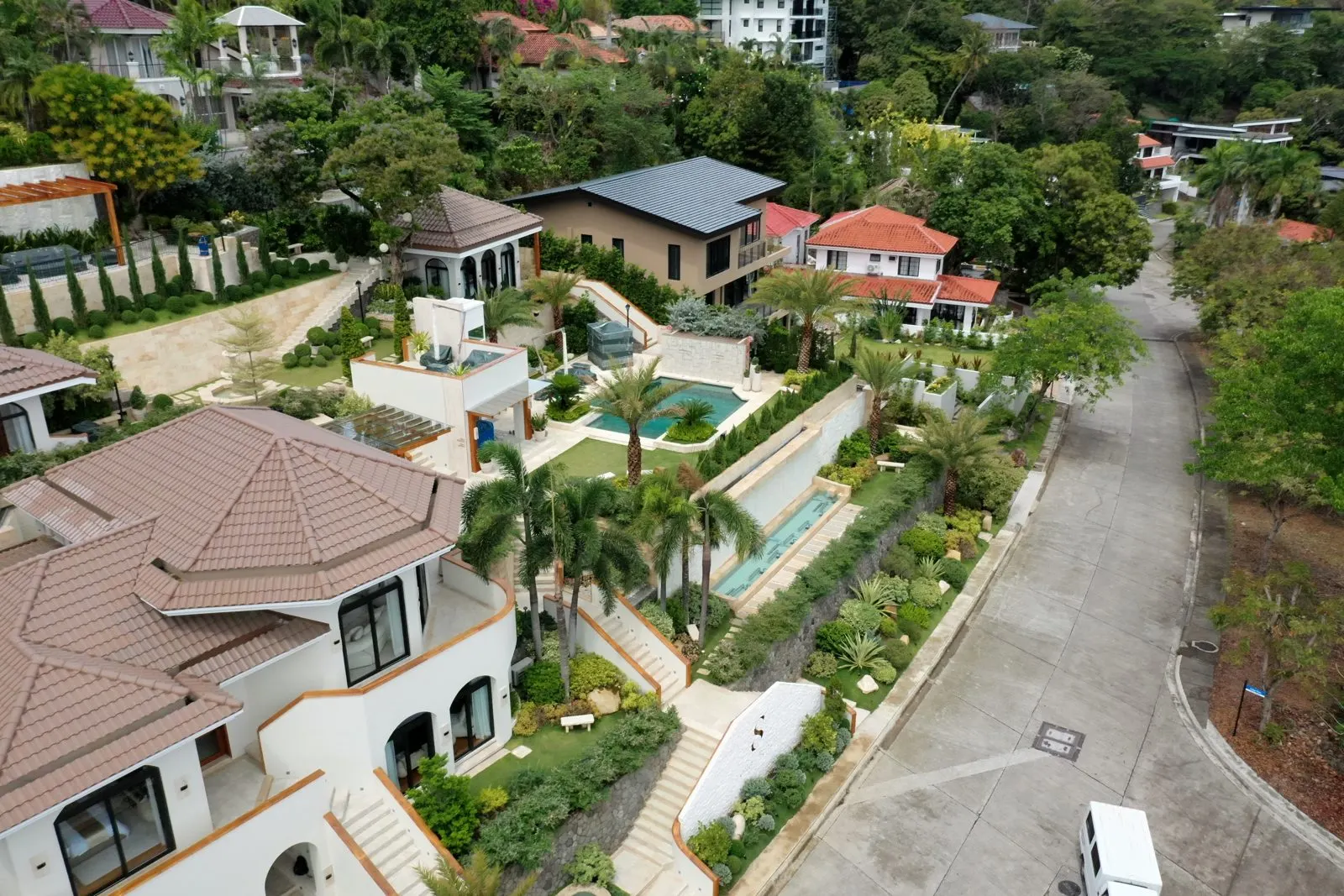 Aerial view of a multi-level hillside coastal estate with terraced lap pools, palm-lined stairways, and full tropical gardens