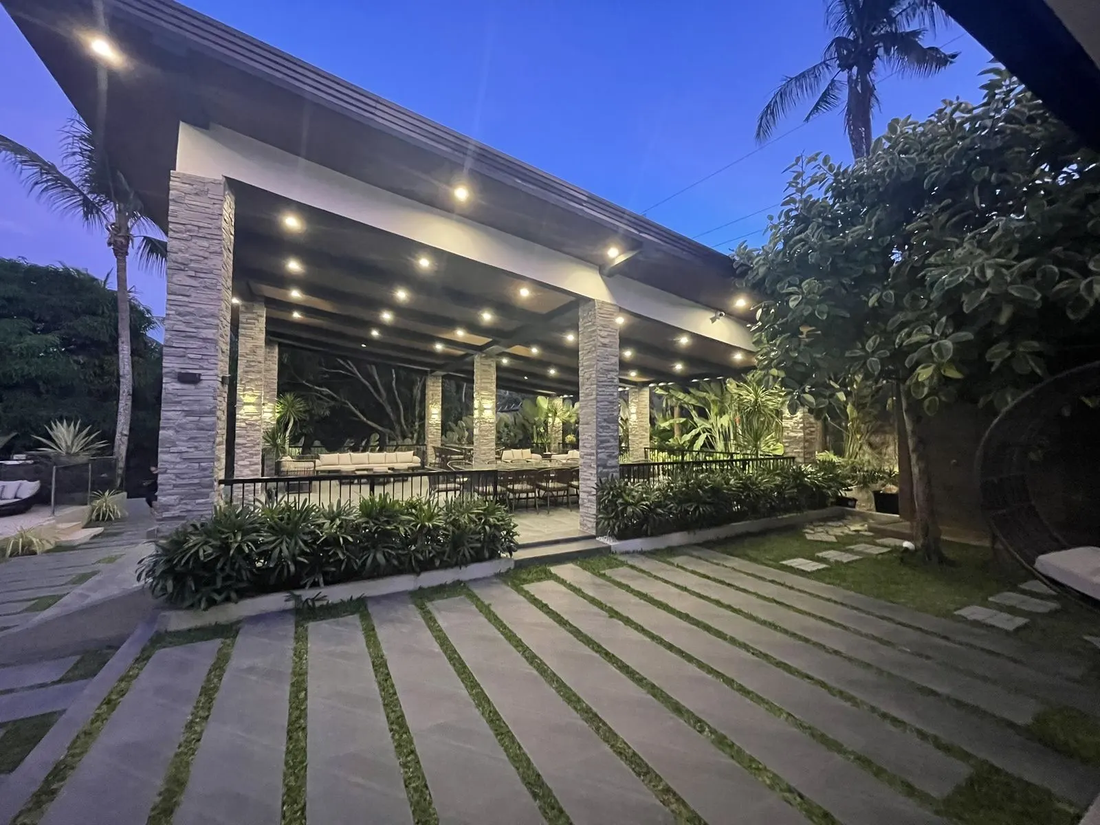 Twilight view of a covered lanai and pool lounge framed by palms and string lighting